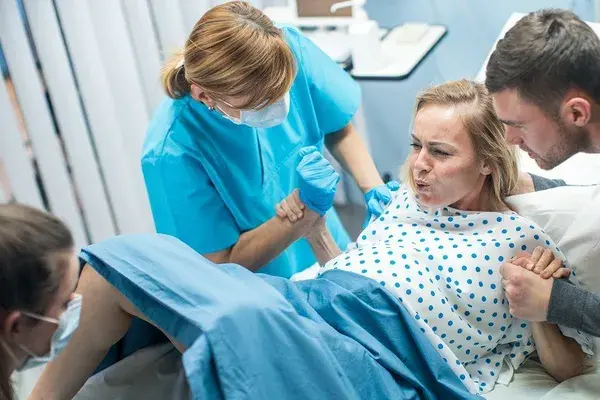 In the Hospital, Woman in Labor Gives Birth, Obstetricians and Doctors Assist, Her Husband Supports Her by Holding Hand. Modern Maternity/ Delivery Ward with Professional Midwives.